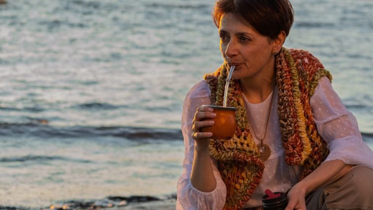 A woman sipping yerba mate while relaxing on a Montevideo beach during sunset.